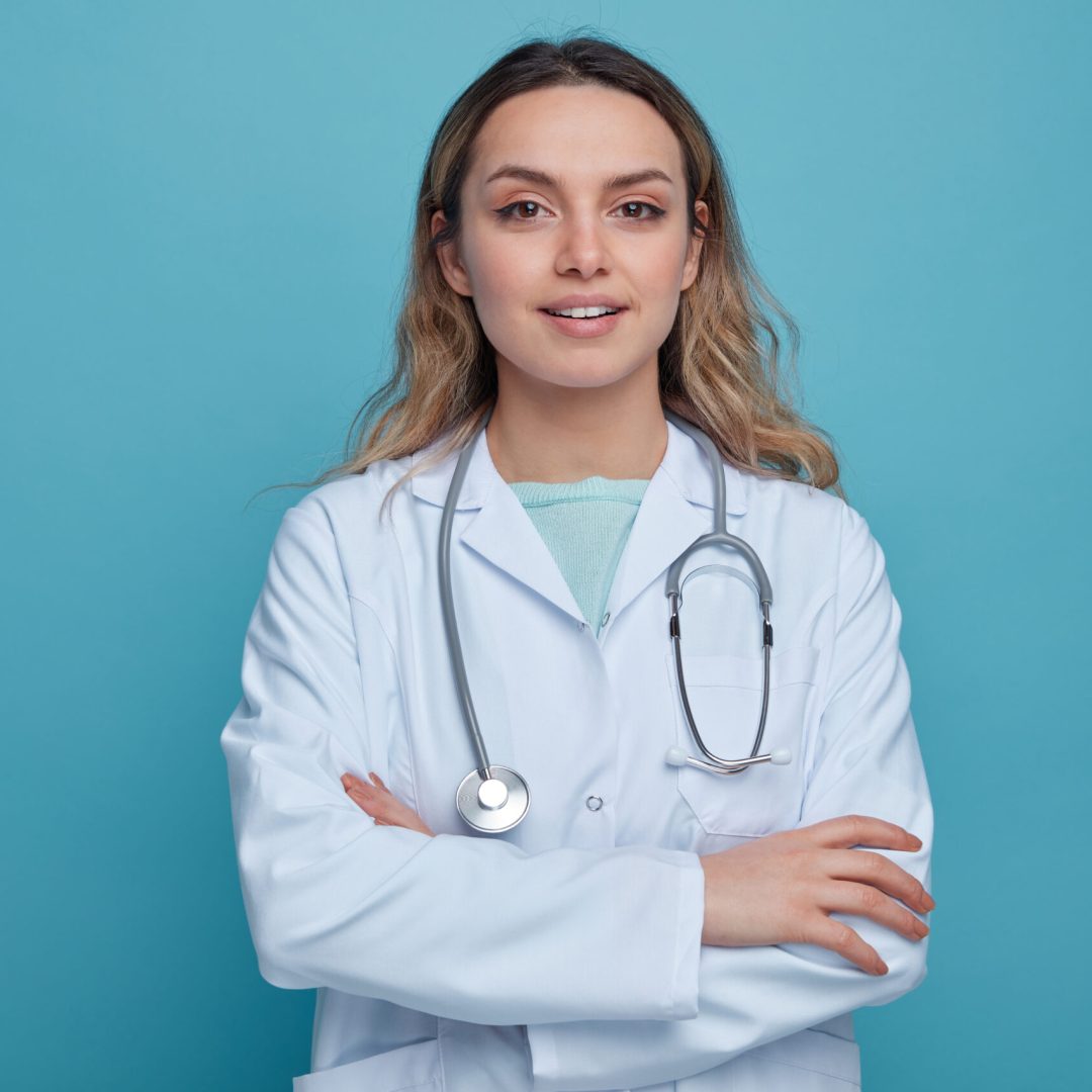 pleased young female doctor wearing medical robe and stethoscope around neck standing with closed posture looking at camera isolated on blue background